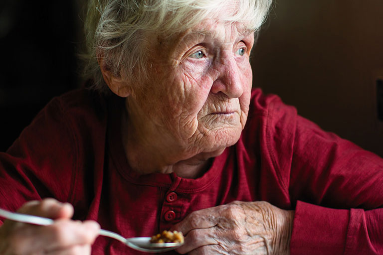 An elderly woman worriedly looking into the distance while eating food.
